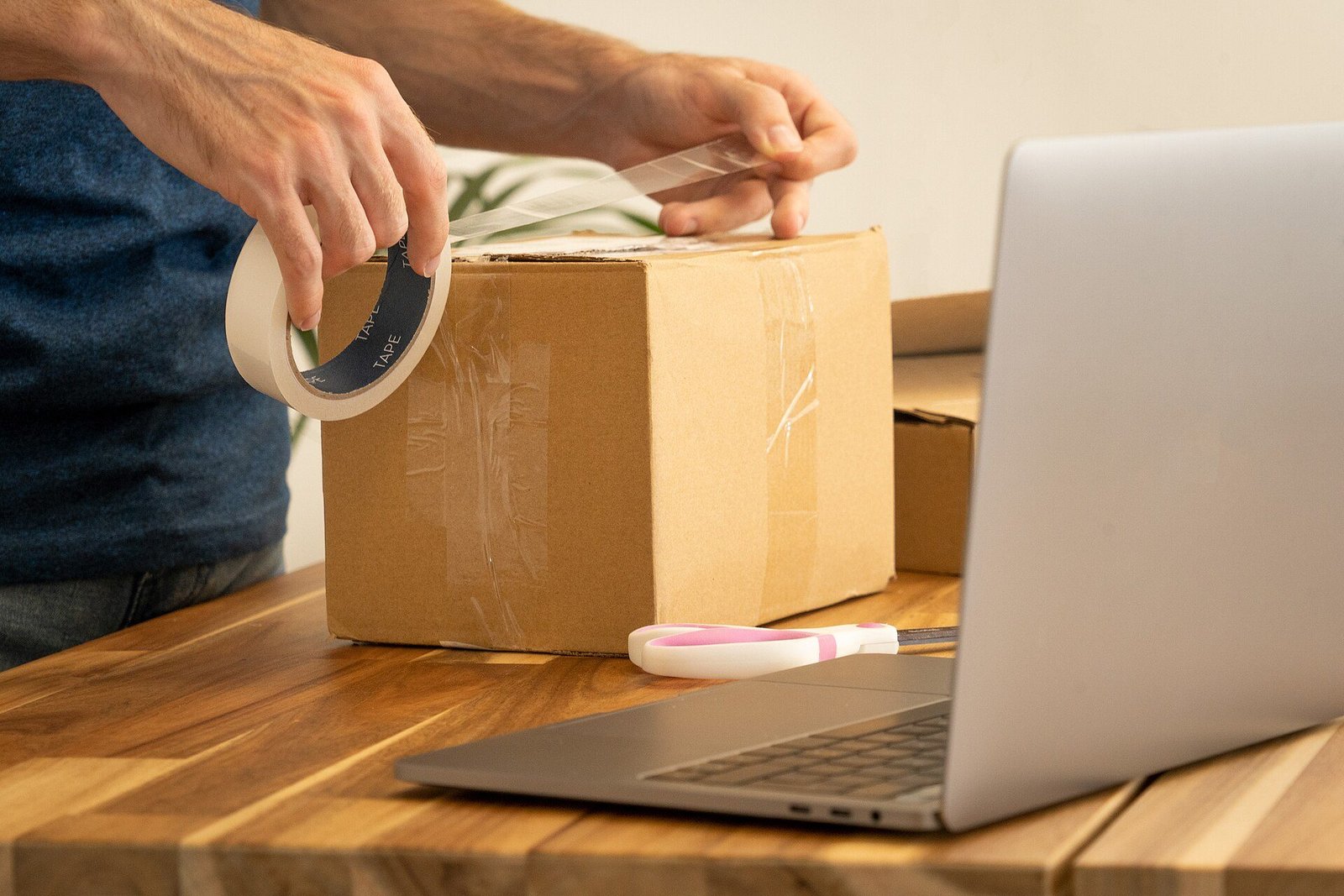 Mains scellant au ruban adhésif un carton d'expédition posé sur une table en bois à côté d'un ordinateur portable ouvert