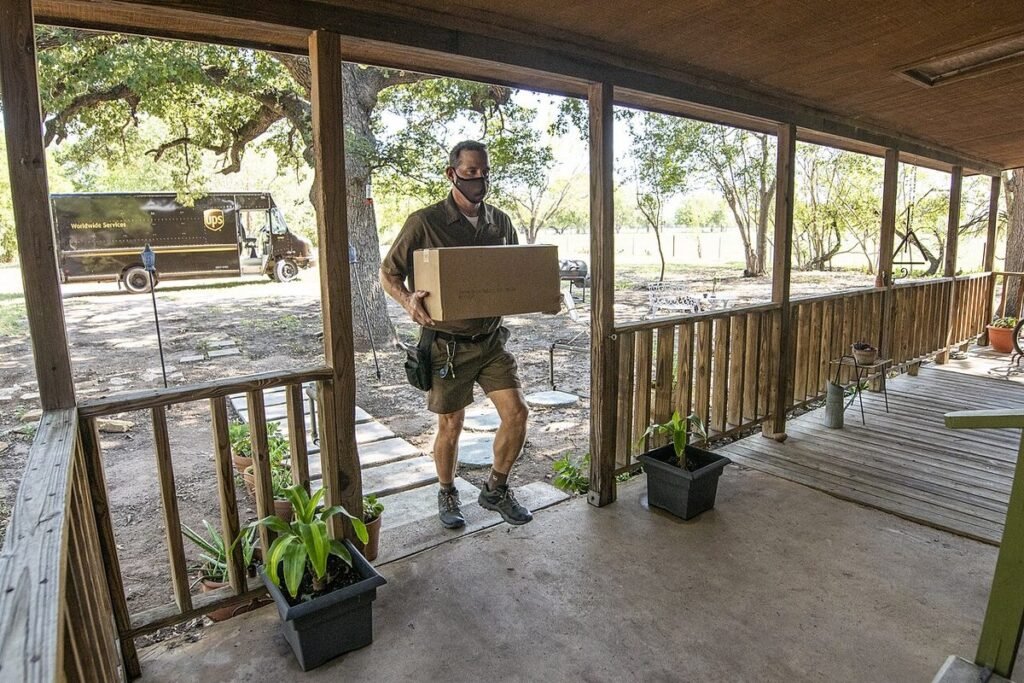 Livreur en uniforme portant un carton jusqu'au porche d'une maison, camionnette de livraison garée derrière lui