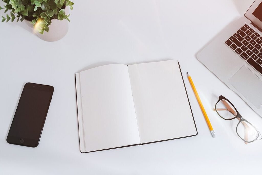 Carnet ouvert, crayon, lunettes, smartphone et ordinateur portable posés sur un bureau blanc